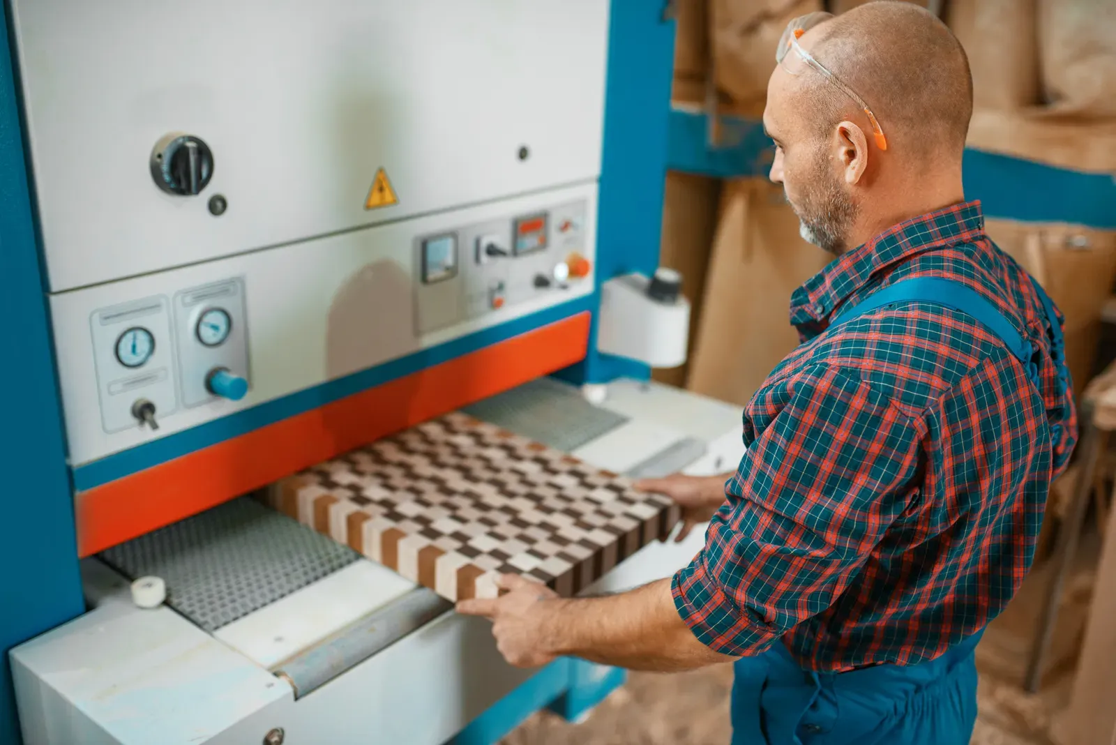 A worker in safety glasses feeds a patterned wood panel into a wide-belt sander.